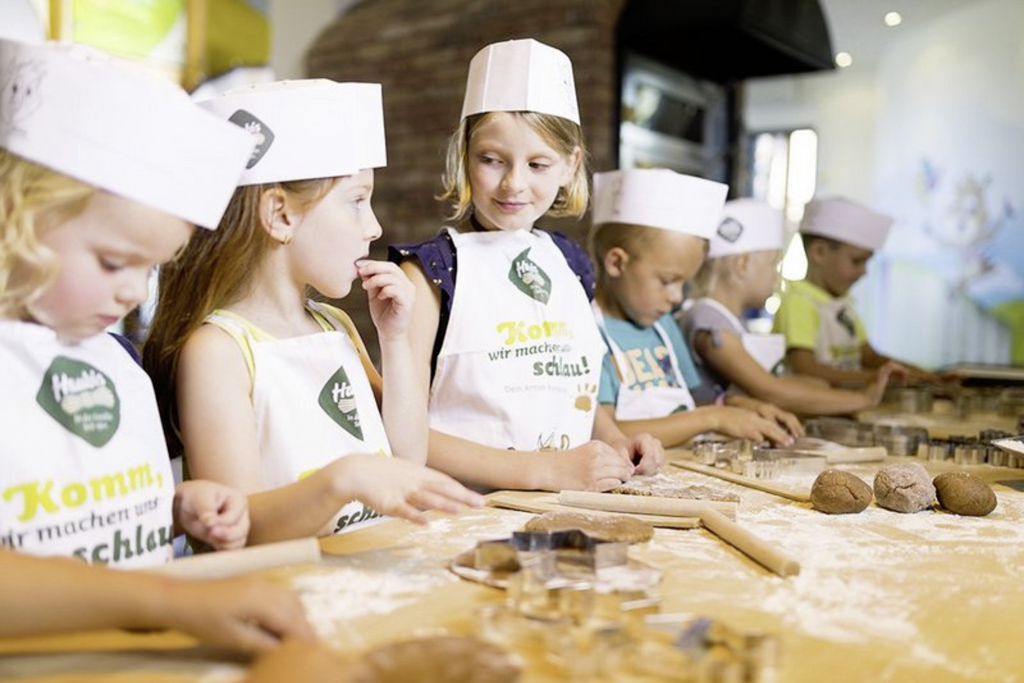 Kinder mit Schürzen und kleinen Kochmützen des Haubiversum stehen lächelnd an einem Tisch mit Teig, Ausstechern und Teigrollen um etwas zu backen.