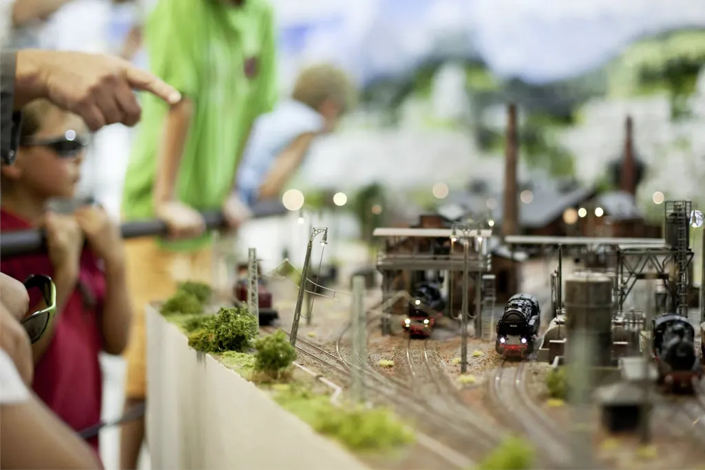Modellbahnhof mit detailgetreuer Landschaft und historischen schwarzen Lokomotiven. An der Seite stehen junge und alte Besucher und bewundern die Ausstellung