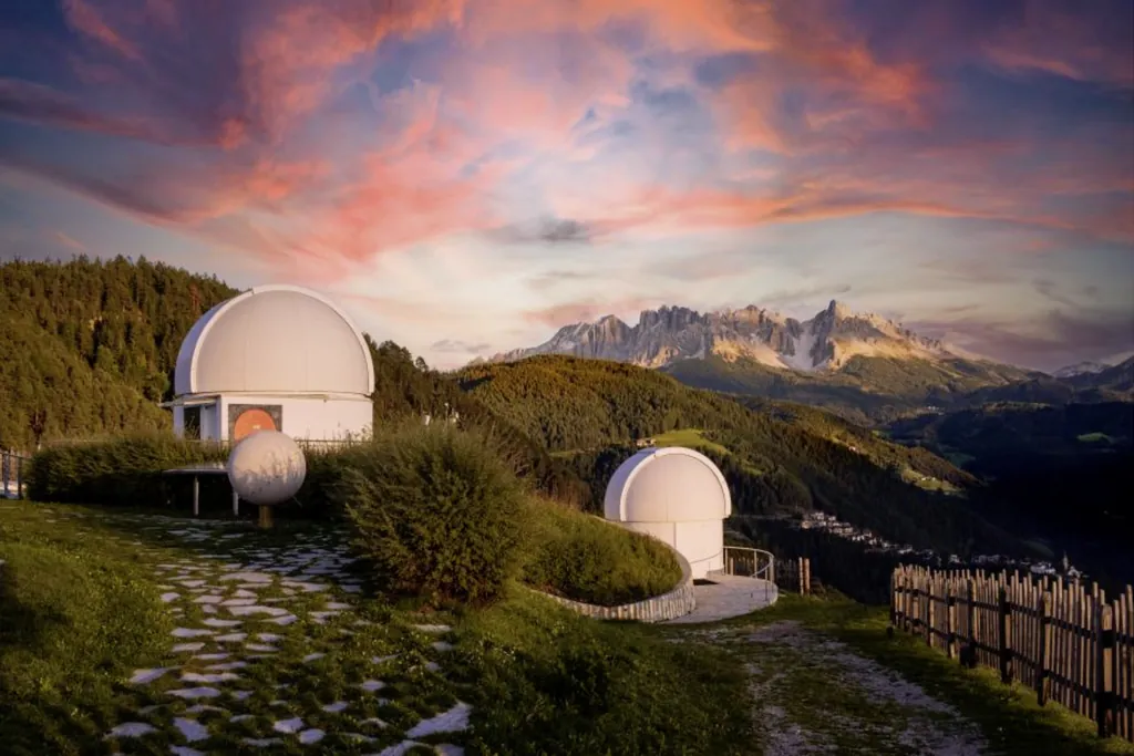 Zwei weiße Kuppeln der Sternwarte Südtirol mitten in der Landschaft. Umgeben von Wiesen, Wäldern und Bergen über denen die Sonne unter geht.