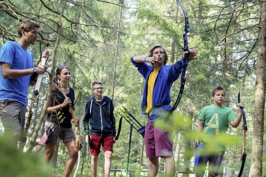 Ein junger Mann spannt gerade Pfeil und Bogen. Um in herum sind Erwachsene und Kinder mit Bogenausrüstung, die ihm genau zusehen.
