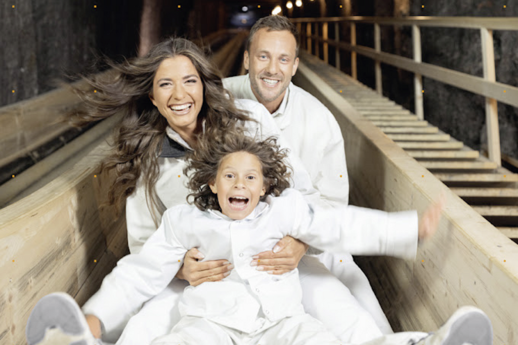 Eine Familie mit Mutter, Vater und Sohn rutscht die Rutsche ins Bergwerk herunter. Die Haare fliegen im Wind. Sie tragen weiße Kittel und Hosen zum Schutz ihrer Kleidung.