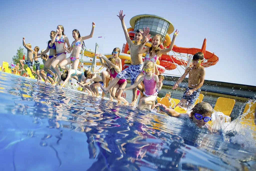 Viele Kinder stehen in einer Reihe am Beckenrand und sind gerade dabei ins Wasser zu springen. Einige haben Schwimmbrillen auf oder halten sich die Nase zu, andere springen mit allen Vieren von sich gestreckt.