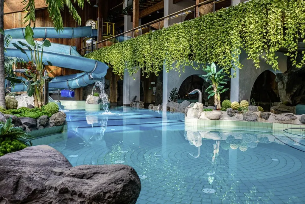 Von der Empore ranken grüne Pflanzen. Eine blaue Rutsche schlängelt sich von oben ihren Weg in das Becken. Rundherum sind große Steine und Pflanzen zur Deko.