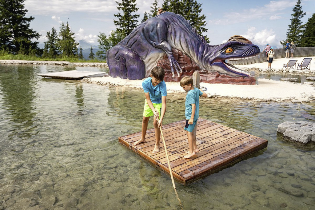 Zwei Jungen stehen auf eine Floß in einem Teich im Hintergrund ist eine riesen große Dinosaurierstatue.