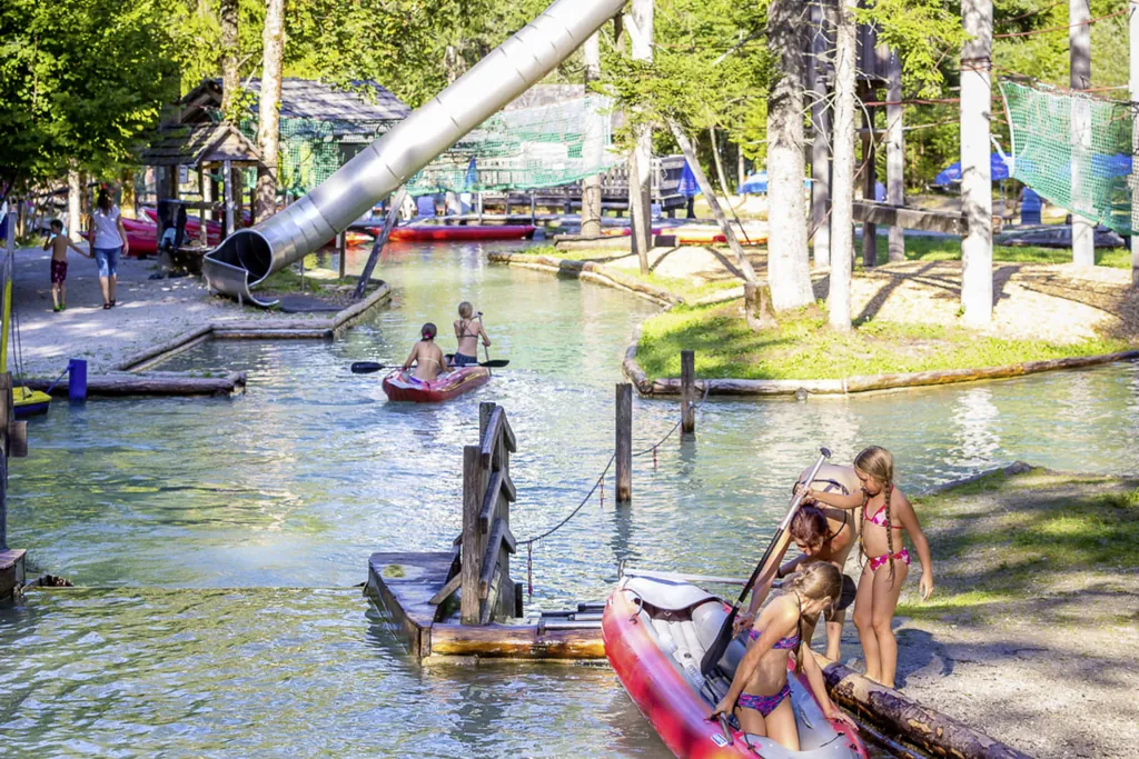 Zwei Mädchen paddeln mit einem aufblasbaren Kanu durch das Wasser. Zwei andere versuchen am Ufer gerade in ein Kanu einzusteigen. Im Hintergrund ist eine Rutsche zu sehen.