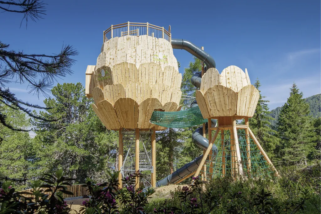 Spielplatz mit Klettergerüst aus Holz in Tannenzapfenform. Man sieht grüne Netze zum Klettern und eine große gewundene Röhrenrutsche.