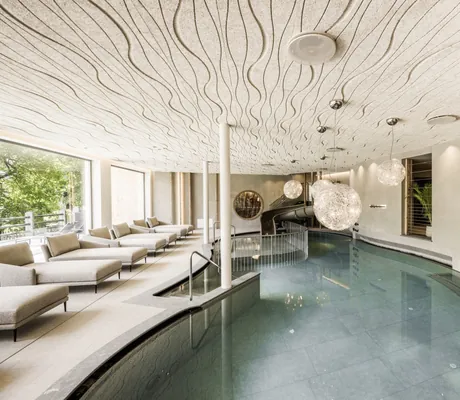geschwungener Indoorpool mit weißen Hängeleuchten und wellenartiger Decke im Excelsior Dolomites Life Resort