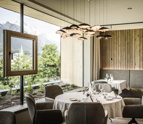Tisch im Restaurant des Excelsior Dolomites Life Resort mit Designer-Hängeleuchte und Fenster im Panoramafenster