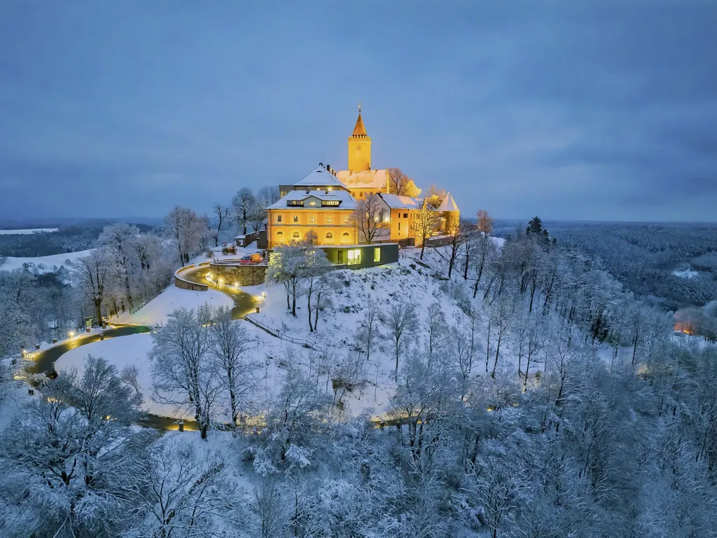 Blick auf die beleuchtete Leuchtenburg am Abend auf einem H&uuml;gel umgeben von vielen B&auml;umen und eine schmale, kurvige Stra&szlig;e, die zur Burg f&uuml;hrt