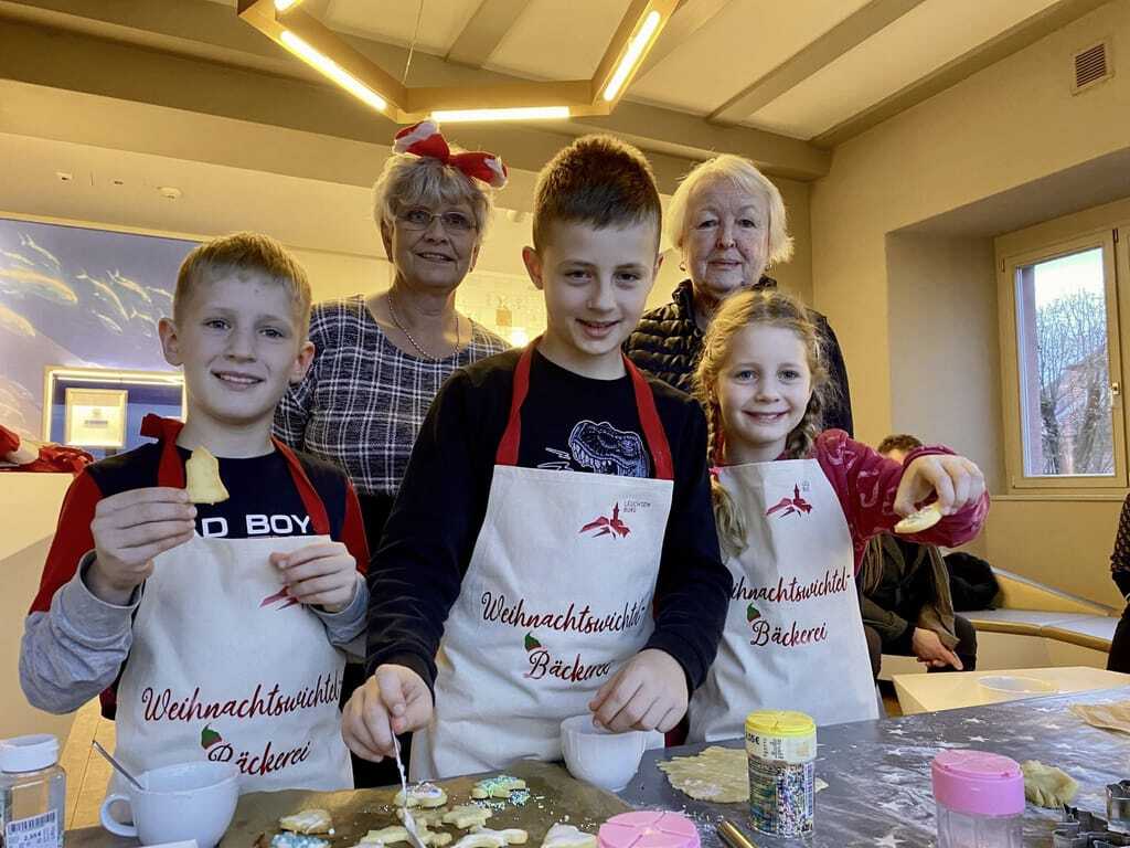 zwei Jungen, ein Mädchen und zwei ältere Frauen lächeln in die Kamera und backen Weihnachtsplätzchen auf der Leuchtenburg