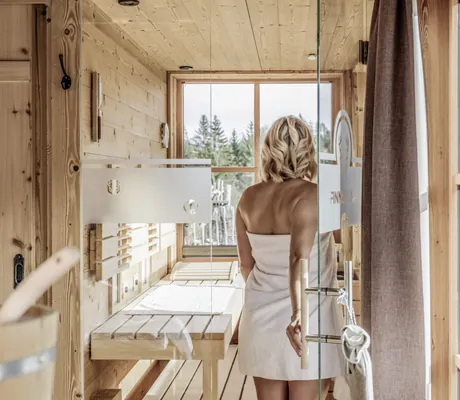 blonde Frau mit weißem Frotteetuch geht in die Sauna im Benglerwald Berg Chaletdorf