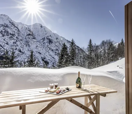 Terrassentisch des Benglerwald - Berg Chaletdorfs gedeckt mit Champagner und Snacks vor meterhohem Schnee, schneebedeckten Bergen und strahlendem Sonnenschein