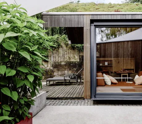 Blick auf und in das Hanghaus aus Holz auf dem Hofgut Hafnerleiten mit gemütlichem Sofa direkt am großen Panoramafenster und Liegestühlen auf der Terrasse
