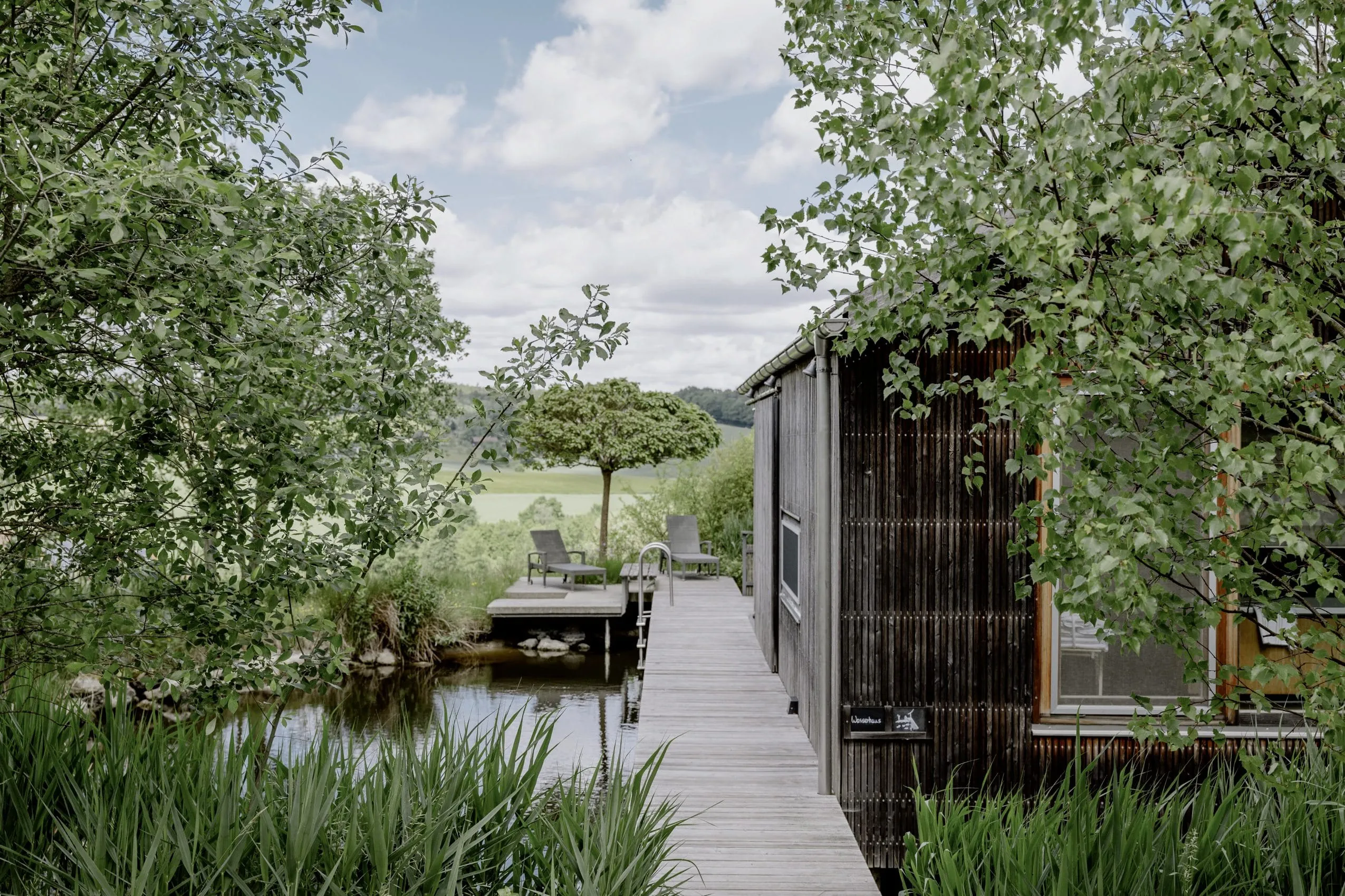 Blick auf das Wasserhaus auf dem Hofgut Hafnerleiten mit Steg, kleinem Teich mit Einstieg und zwei Liegestühlen