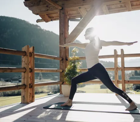 Frau mit weißem Top und schwarzer Hose macht Yoga auf einer Terrasse in den Chalets Valsegg