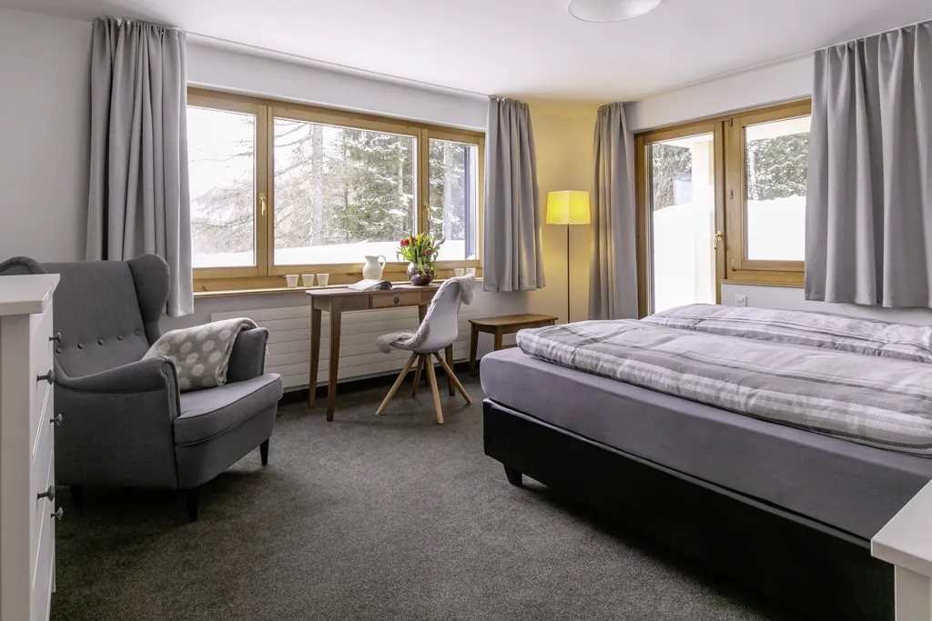 Schlafzimmer des Chalet vue au lac mit Doppelbett, grauem Sessel. kleinem Holztisch mit weißem Stuhl und vielen Fenstern