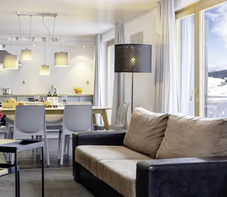 lichtdurchfluteter Wohn-/Essbereich im Chalet vue au lac mit kleinem Sofa, großer schwarzer Stehlampe und Esstisch und Küche im Hintergrund