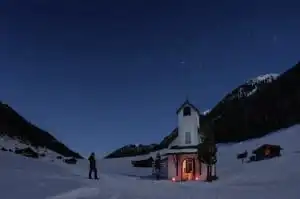 Person steht neben einer beleuchteten Kapelle in verschneiter Berglandschaft unter sternenklarem Nachthimmel in der Nähe der Summit Lodges