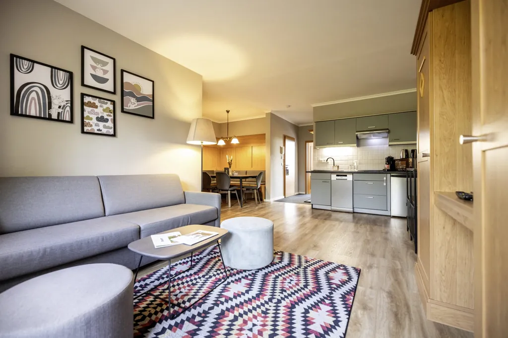 Blick in die Premium Plus Suite des Familotel Elldus Resort mit grauem Sofa, gemustertem Teppich, Küche und Essbereich im Hintergrund