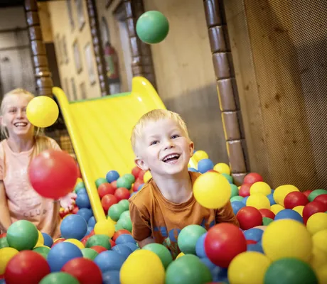 Blondes Mädchen und blonder Junge spielen lachend im bunten Bällebad des Familotel Elldus Resort mit gelber Rutsche im Hintergrund