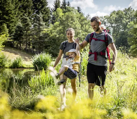 Eltern lachen und halten kleines Mädchen, das in die Luft springt an den Händen während einer Wanderung in grüner Umgebung in der Nähe des Familotel Elldus Resort