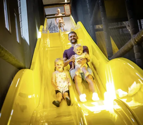 Junger Mann rutscht mit zwei kleinen Mädchen eine gelbe Rutsche im Familotel Elldus Resort hinunter