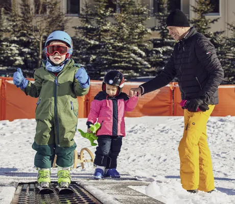 Kleiner Junge auf Skiern in Skibekleidung auf einem schneebedeckten Förderband direkt neben dem Familotel Elldus Resort und kleines Mädchen im Hintergrund hält die Hand eines Mannes.