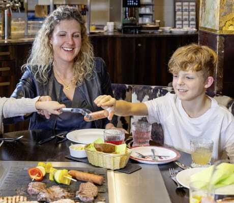 blonde Frau mit Locken sitzt mit zwei Kindern am Tischgrill im Feldberger Hof und grillt Fleisch