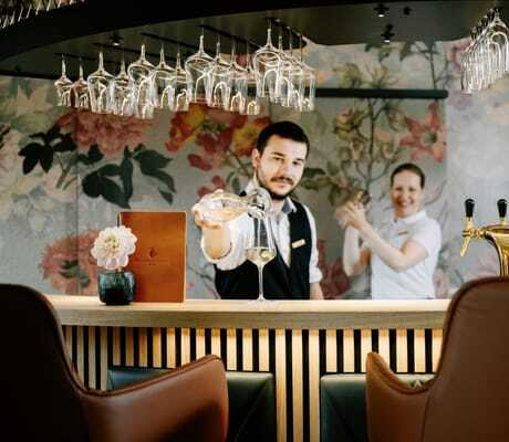 Barkeeper schenkt Wein aus Karaffe in ein Weinglas an der Bar des Familotel Landhaus Averbeck ein, Barkeeperin mit Cocktailshaker im Hintergrund
