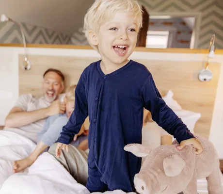 kleiner, blonder Junge steht lachend mit einem Schweinchen-Kuscheltier auf einem Doppelbett des Familotel Landhaus Averbeck, im Hintergrund die restliche Familie lachend