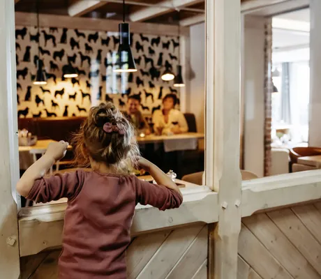 Mädchen steht mit dem Rücken zur Kamera und schaut durch ein Fenster in den Restaurantbereich des Familotel Landhaus Averbeck