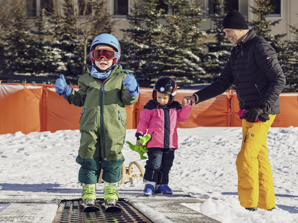 Kleiner Junge auf Skiern in Skibekleidung auf einem schneebedeckten Förderband direkt neben dem Familotel Elldus Resort und kleines Mädchen im Hintergrund hält die Hand eines Mannes.