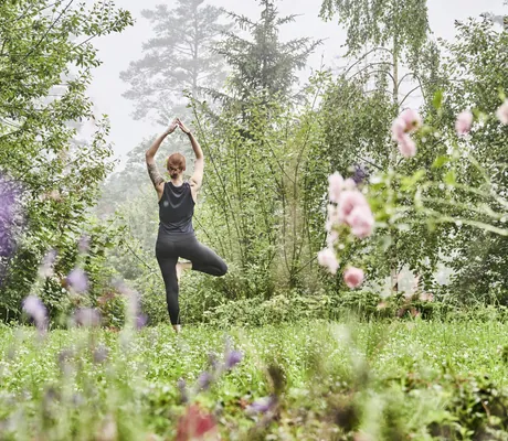 Frau in schwarzer Sportkleidung macht eine Yogaübung inmitten grüner, blühender Natur in der Nähe des Hotel Kronelamm