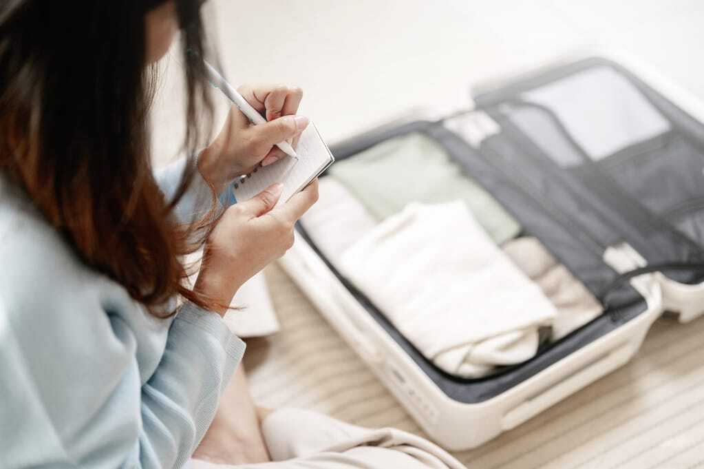 Junge Frau packt den Koffer für ihren Wellnessurlaub und schreibst das Wichtigste auf einem kleinen Block