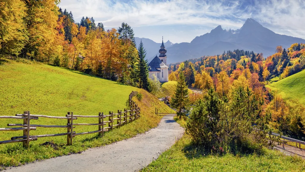 Herbstlandschaft in Berchtesgaden mit gelb, orange und rotgefärbten Bäumen, einer kleiner Kirche und Berggipfeln im Hintergrund