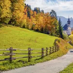 Herbstlandschaft in Berchtesgaden mit gelb, orange und rotgefärbten Bäumen, einer kleiner Kirche und Berggipfeln im Hintergrund