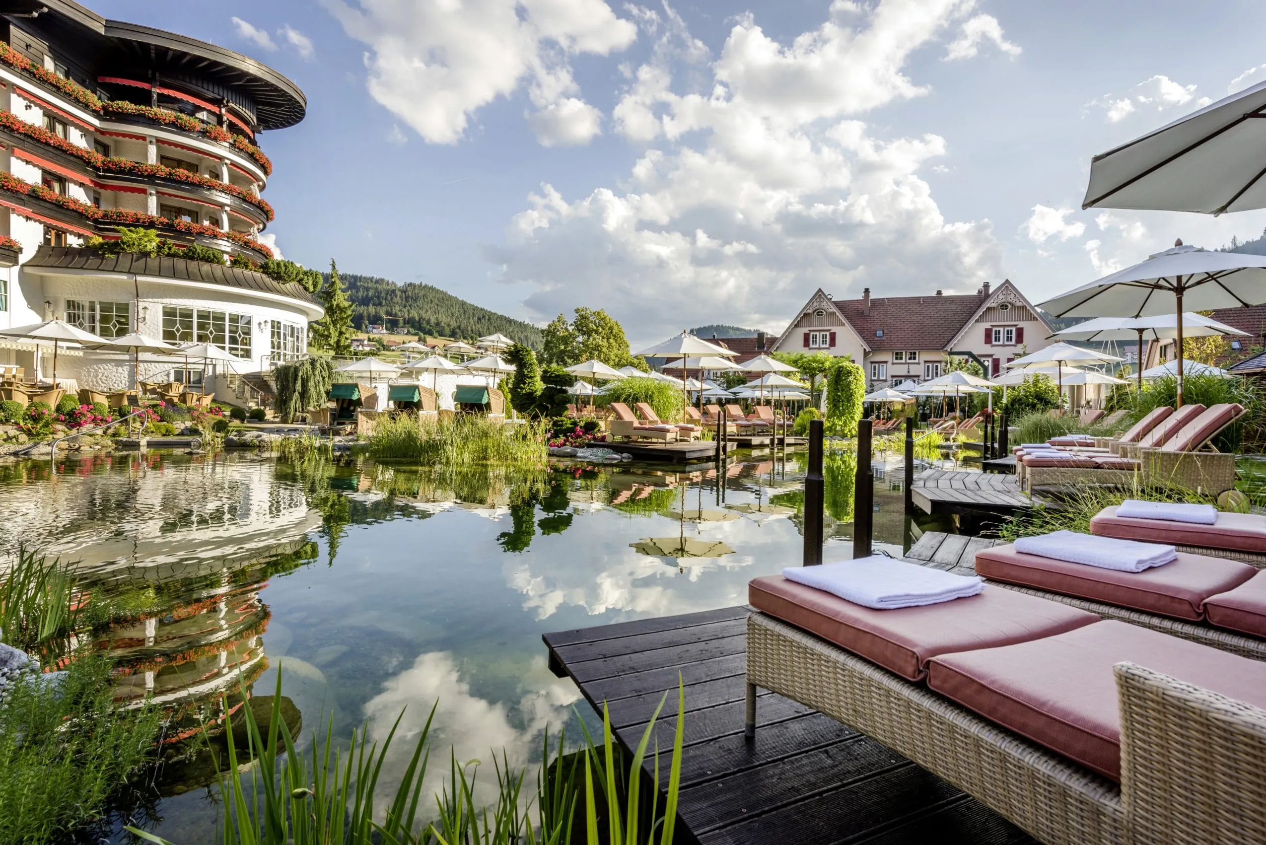 Außenansicht des Hotel Bareiss mit Naturteich im Vordergrund und einigen Sonnenliegen sowie weißen Sonnenschirmen