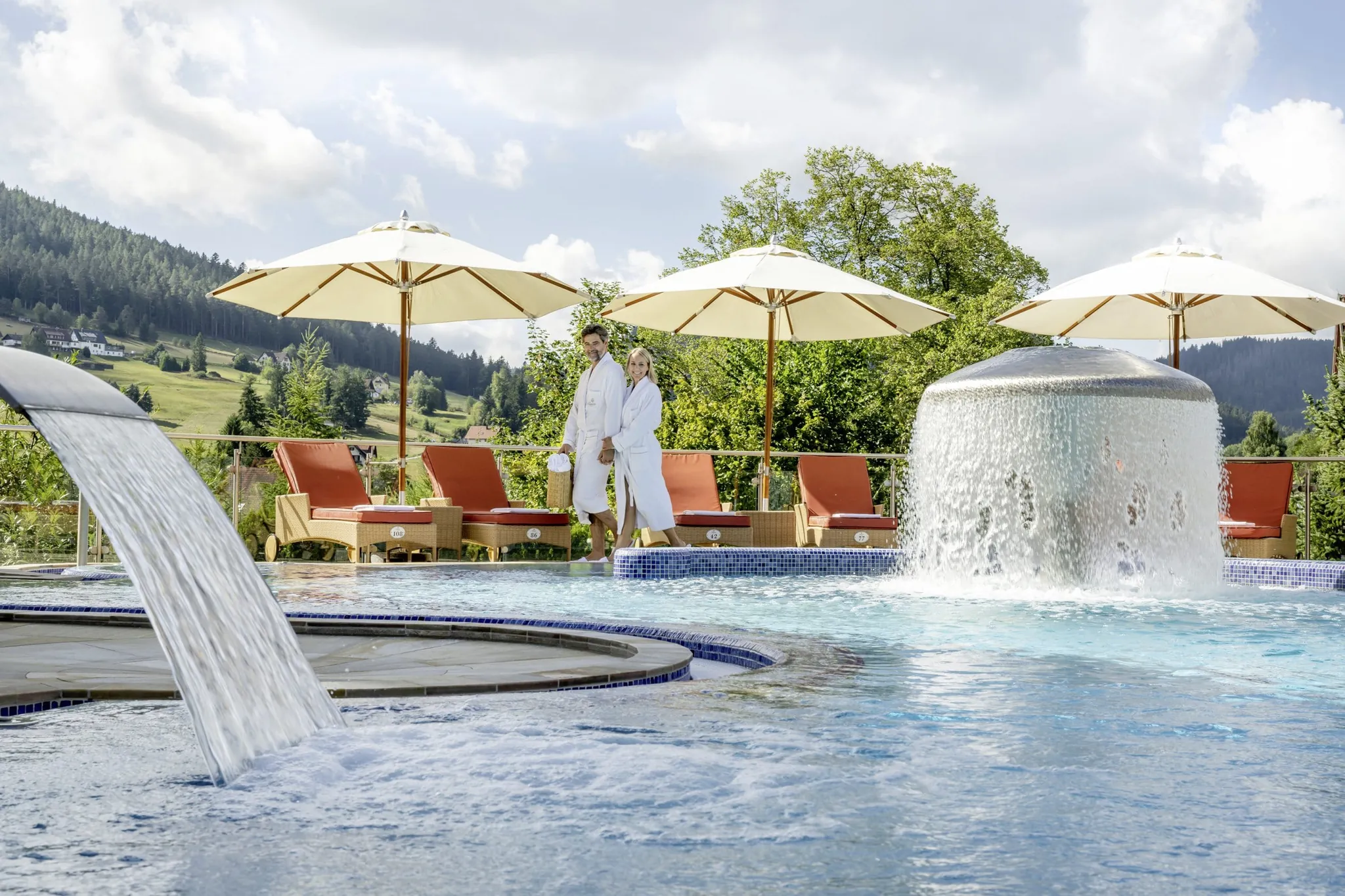 Pärchen in weißen Bademänteln läuft am Outdoorpool des Hotel Bareiss entlang, Sonnenliegen und -schirme im Hintergrund