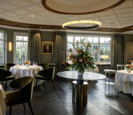 modernes Restaurant des Hotel Bareiss mit runden, mit weißen Tischdecken überzogenen Tischen, khakifarbenen Samtstühlen und großem Blumenstrauß in der Mitte