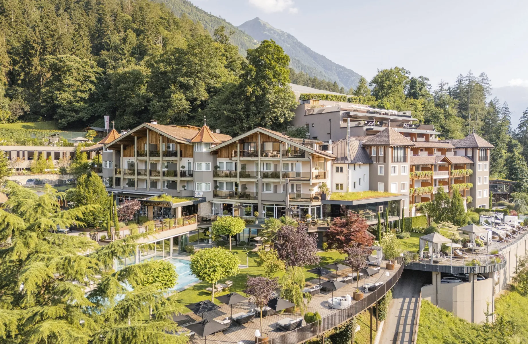 Außenansicht des Alpenschlössel mit einigen Bäumen, Outdoorpool, Liegemöglichkeiten und Wald und Berge im Hintergrund