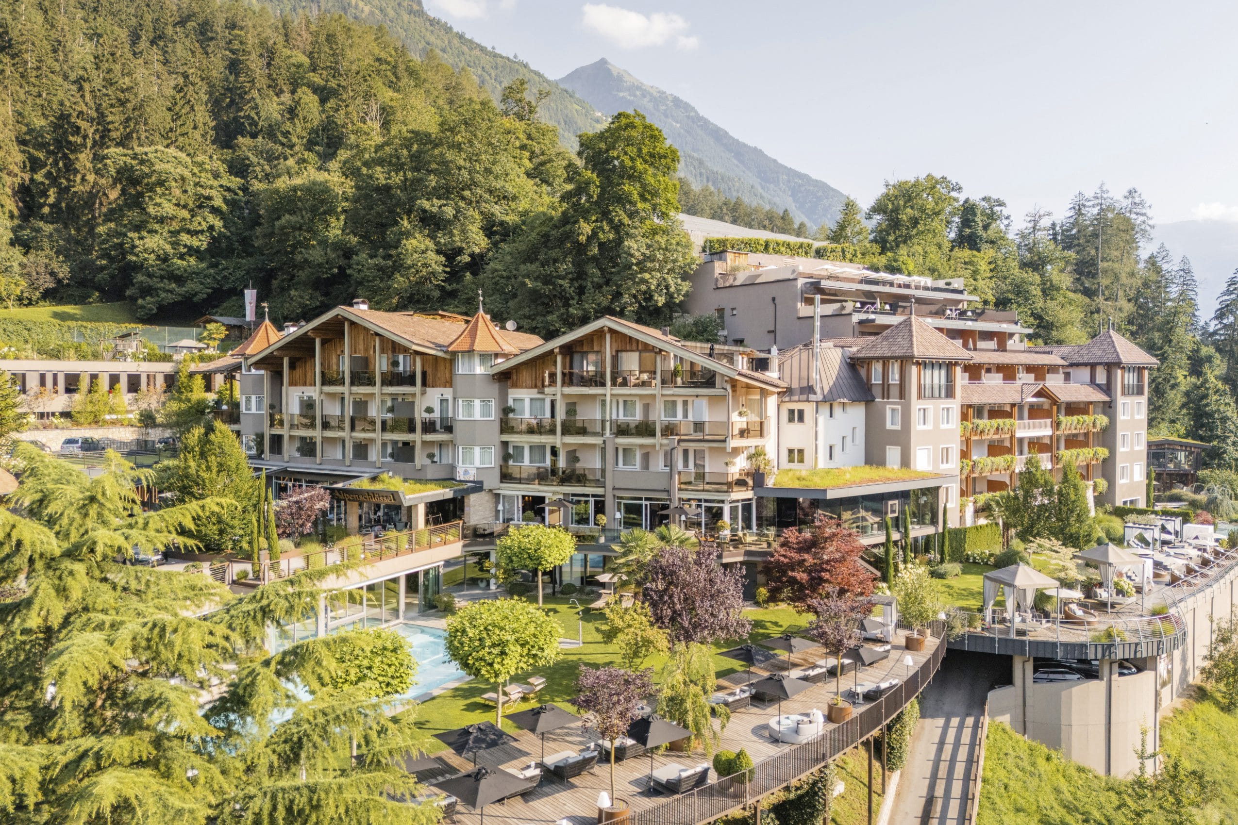 Außenansicht des Alpenschlössel mit einigen Bäumen, Outdoorpool, Liegemöglichkeiten und Wald und Berge im Hintergrund