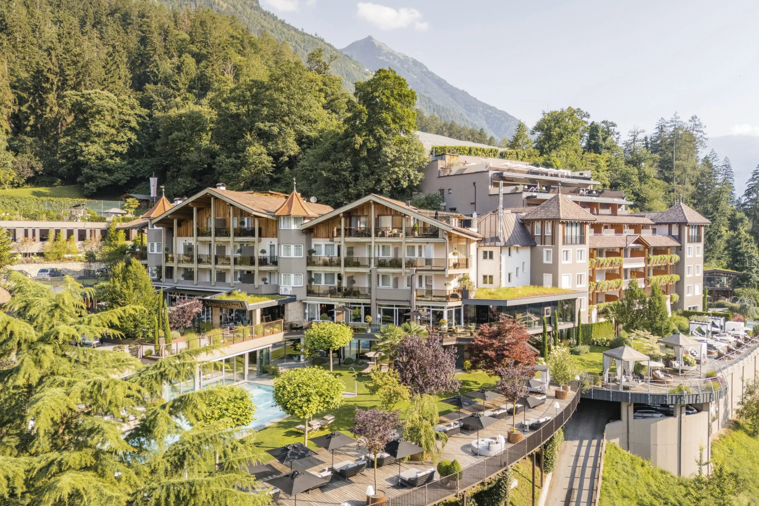 Außenansicht des Alpenschlössel mit einigen Bäumen, Outdoorpool, Liegemöglichkeiten und Wald und Berge im Hintergrund