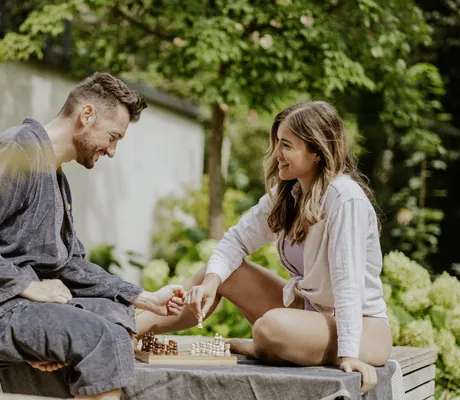 Mann in Bademantel udn Frau in Bluse sitzen auf Holzponton des Klosterhof - Alpine Hideaway & Spa und spielen Schach