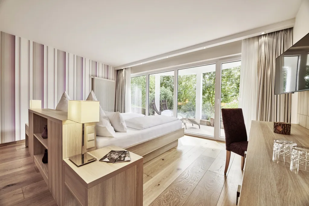 Blick in ein lichtdurchflutetes, helles Zimmer des Wellnesshotel Kronelamm mit großen Fenstern, Holzeinrichtung, Doppelbett, TV und Holzboden