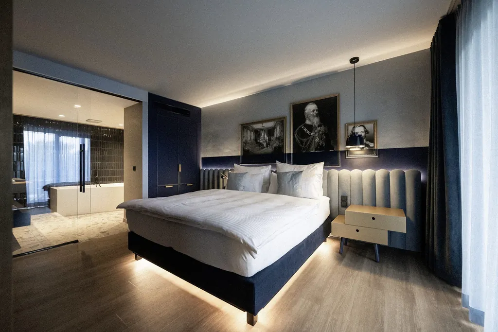 Blick in die dunkelblau und grau eingerichtete Suite Bertl des Wellnesshotel Wittelsbach mit großem Doppelbett, historischen Gemälden an der Wand und verglaste Tür mit Blick ins Bad mit Badewanne