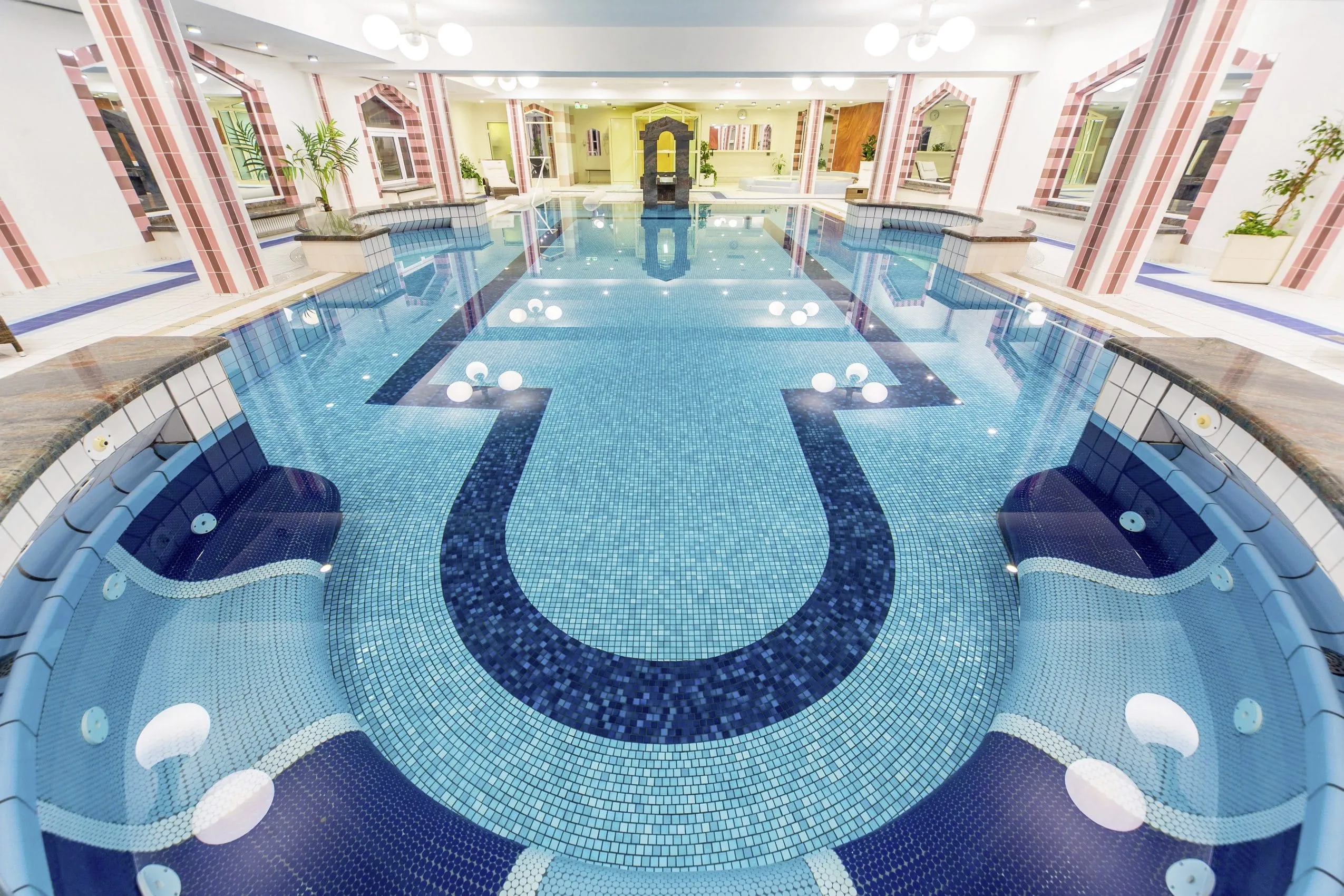 Eleganter Indoor-Pool mit aufwendigen Mosaikfliesen in verschiedenen Blautönen und integrierten Massageliegen, umgeben von stilvoll gestalteten Säulen und Bögen.