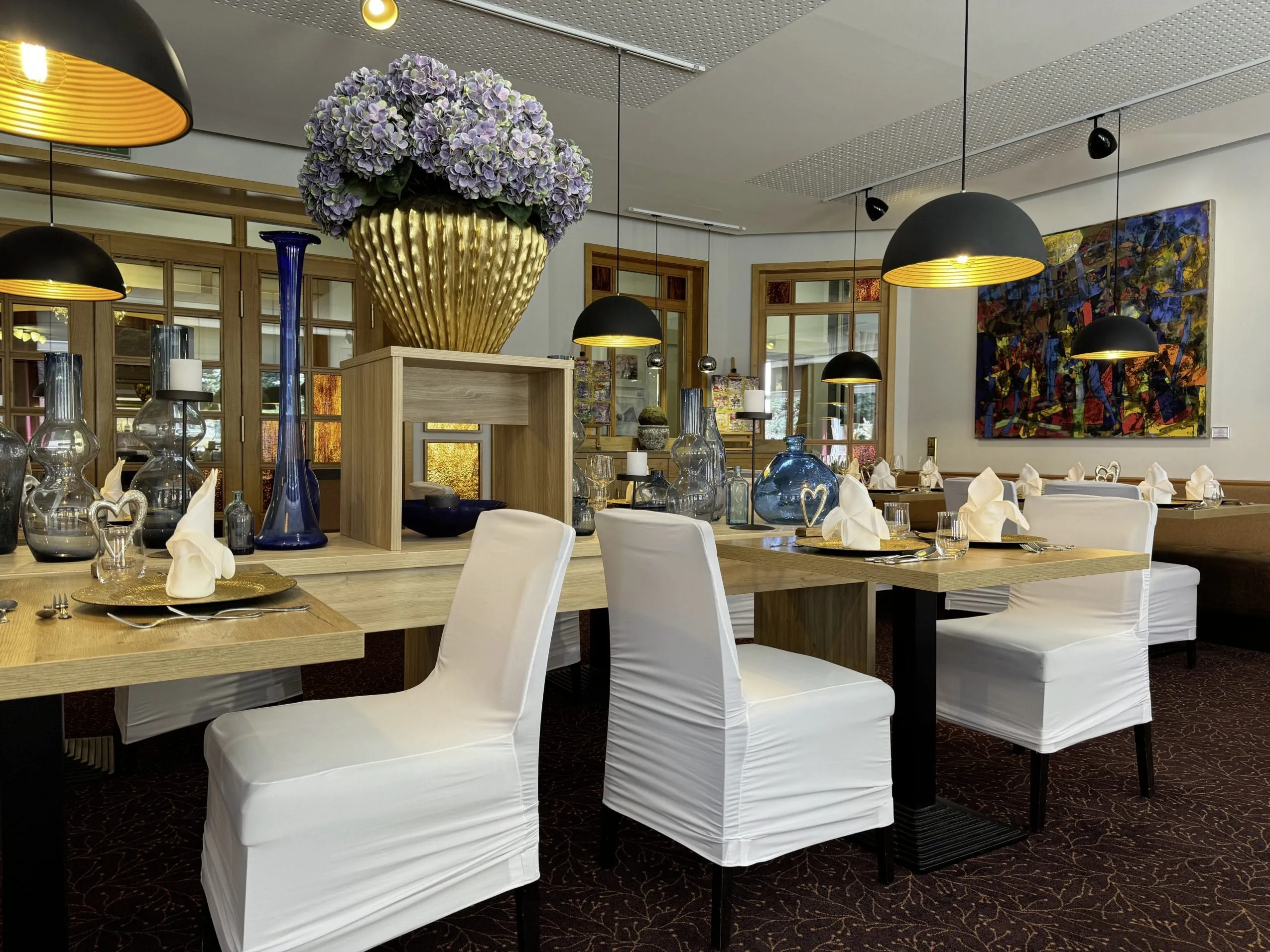 Restaurant mit elegant gedeckten Tischen, weißen Stuhlhussen, dekorativen Glasvasen und einem großen Blumenarrangement in einer goldenen Vase im Zentrum.