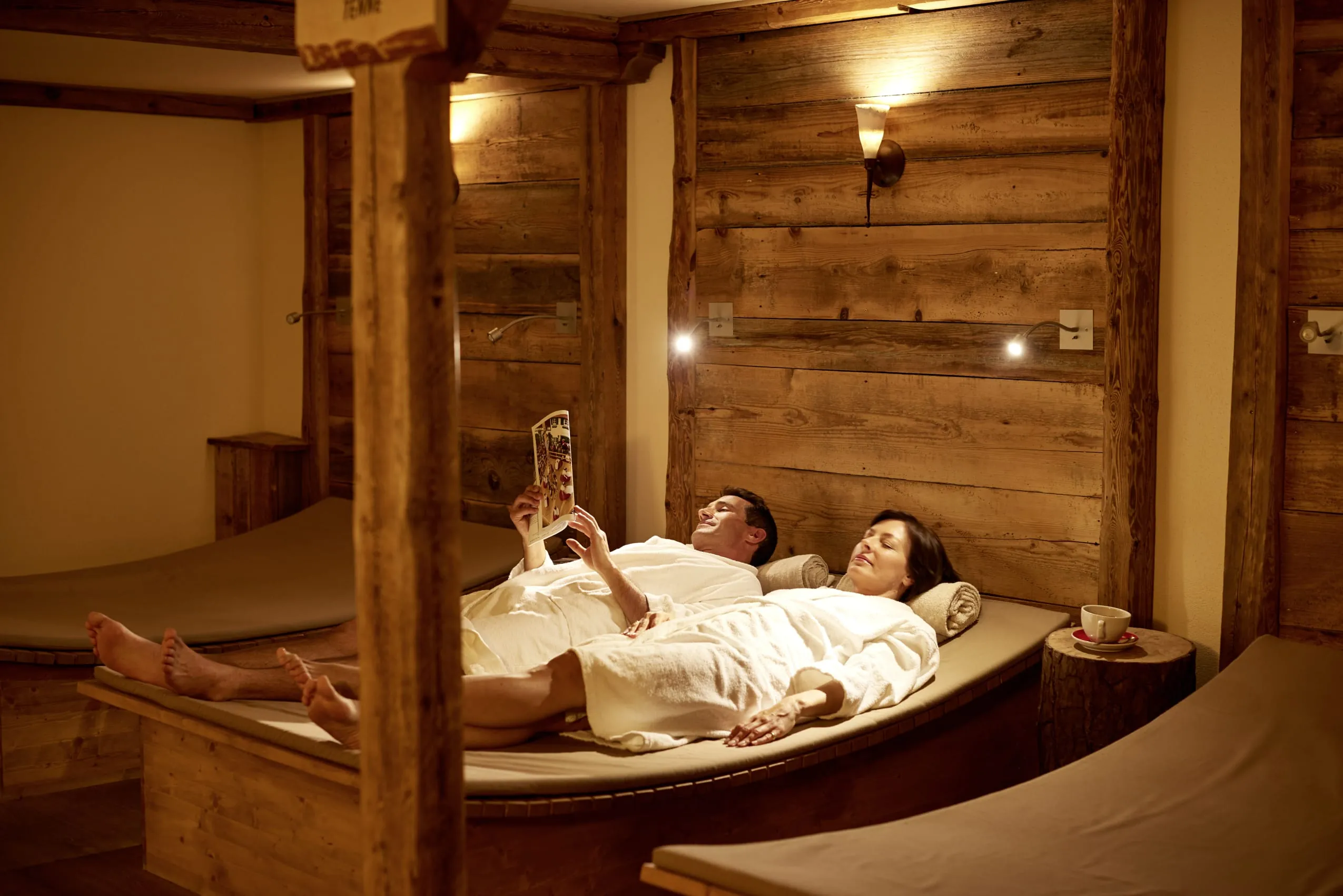 Ein Pärchen in weißen Bademänteln entspannt im rustikal gestalteten, dem Allgäu nachempfundenen Ruhebereich der Saunalandschaft mit vielen Holzelementen und gemütlichen Day-Beds.