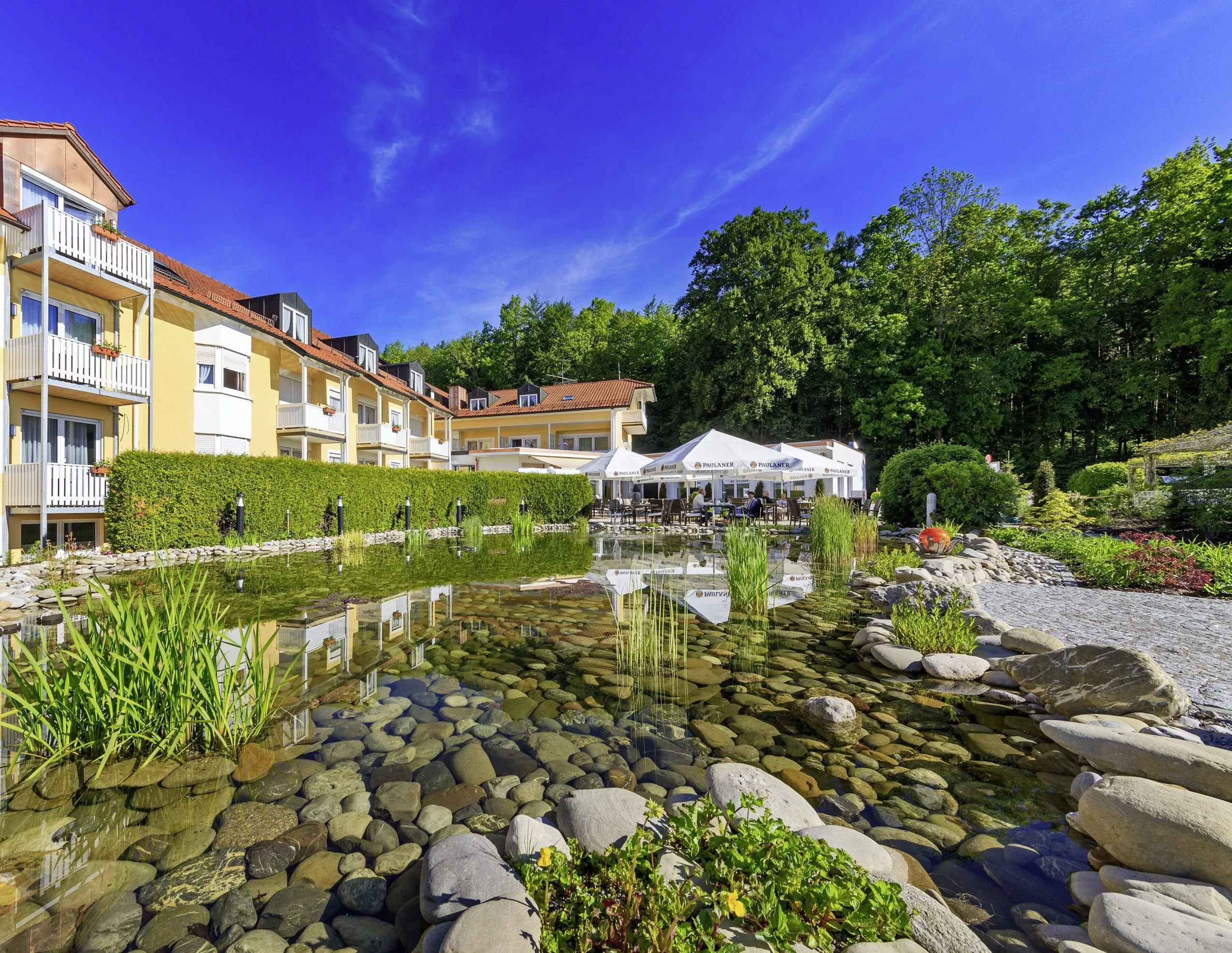 Außenansicht des Hotels mit seiner dezent orangefarbenen Fassade und weißen Balkons mit dem davor liegenden Naturteich.