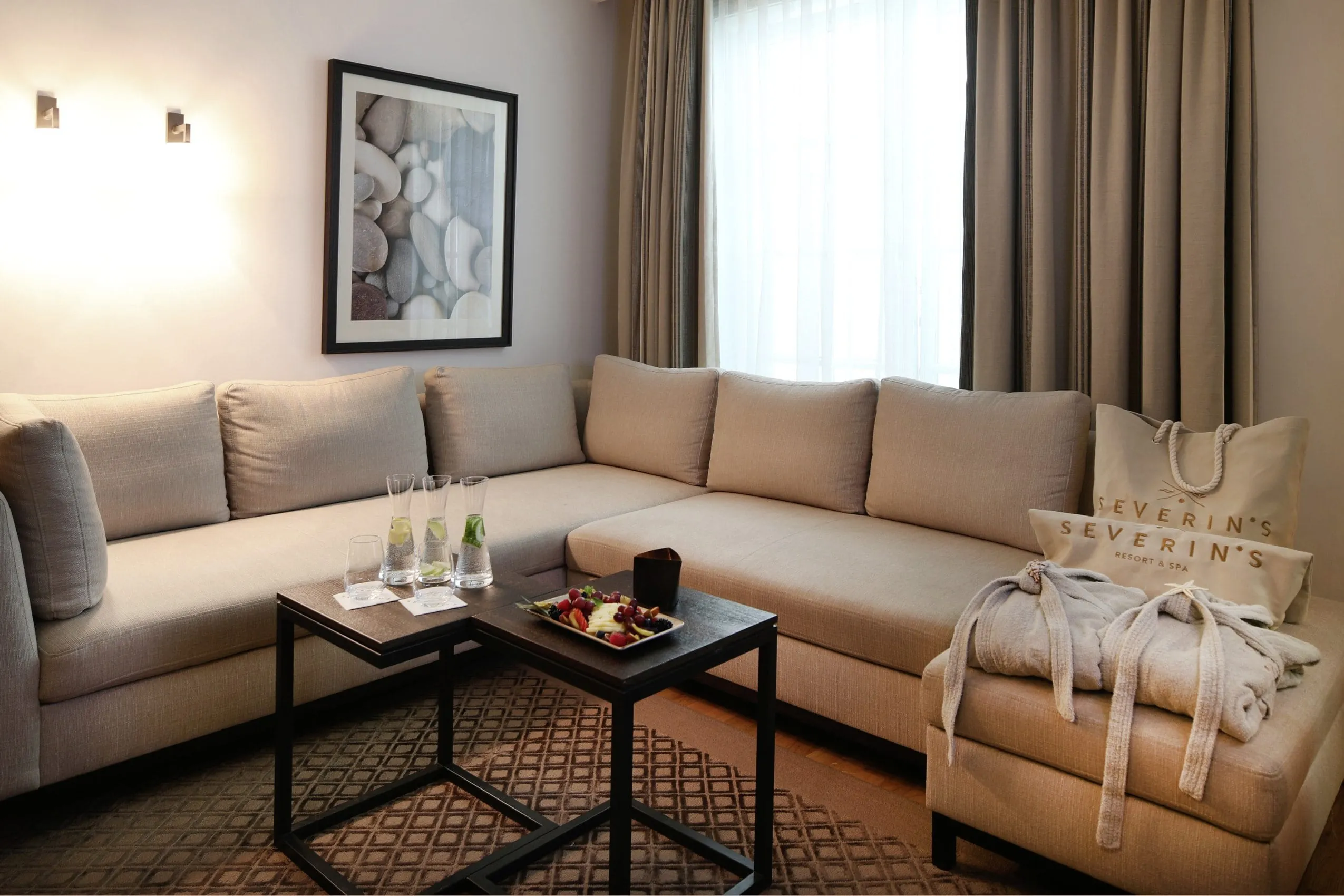 Wohnbereich de Day Spa Suite mit grauem Sofa auf dem Badem&auml;ntel und Spataschen bereitliegen. An der Wand hinter dem Sofa h&auml;ngt ein Bild und die Vorh&auml;nge sind farblich auf das Sofa und den Teppich davor abgestimmt.